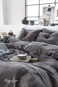 an unmade bed with grey linens in a white room next to a window