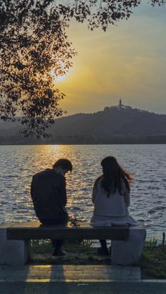 two people are sitting on a bench near the water at sunset or sunrise, looking out to sea