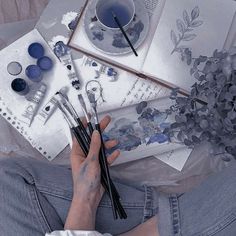 a person sitting on the floor with some paint and brushes in their hands next to an open book