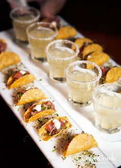 a white tray topped with lots of tacos and glasses filled with drinks on top of it