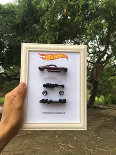 a person holding up a framed photo with three toy cars in front of trees and grass