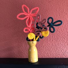 a yellow vase with crocheted flowers in it on a shelf against a pink wall