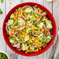 a red bowl filled with corn, cucumber and onions on top of a white table