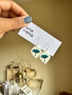 a pair of white and green earrings with leaves on them are being held up by a woman's hand