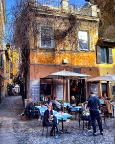 people sitting at tables in an alleyway with buildings and trees on the other side