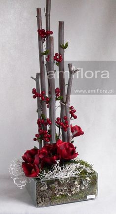 red flowers and branches are arranged in a square vase on a white surface with moss