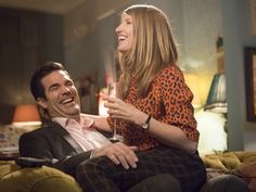 a man and woman sitting on a couch holding wine glasses in their hands, laughing