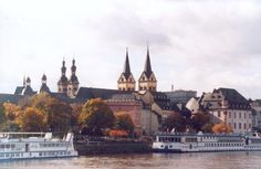 two large boats on the water in front of some buildings and trees with fall colors