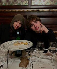 two people sitting at a table with food and wine glasses on it, one person is holding his head