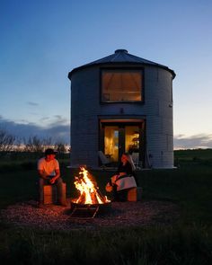 two people sitting around a fire pit in front of a round house at night with the lights on