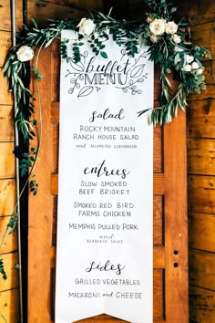 a menu for a wedding with greenery and white flowers on it, hanging from a wooden wall