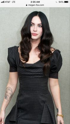 a woman with long dark hair wearing a black dress and posing for the camera in front of a wall