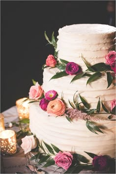 a white wedding cake with pink and purple flowers on the top, surrounded by candles