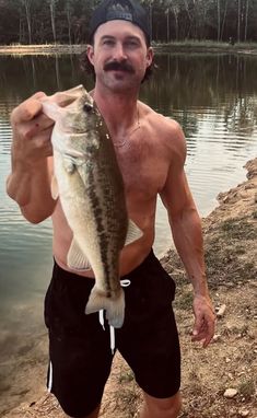 a shirtless man holding a large fish in his right hand while standing next to a body of water