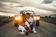 a man and woman sitting on the ground next to a red car with headlights on