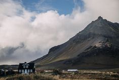 a house sits in the middle of a field with mountains in the background and clouds overhead