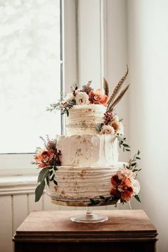 a white wedding cake with feathers and flowers on the top is sitting on a table in front of a window