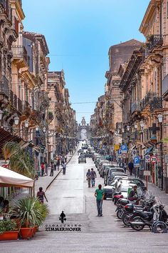 people are walking down an empty street lined with parked cars and motor scooters