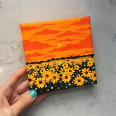 a hand is holding up a small painting with sunflowers in the foreground