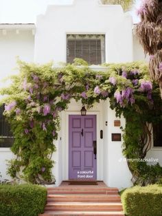 a purple front door is surrounded by greenery and bushes on the side of a white house