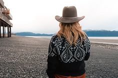 a woman wearing a cowboy hat walks on the beach near a building with mountains in the background