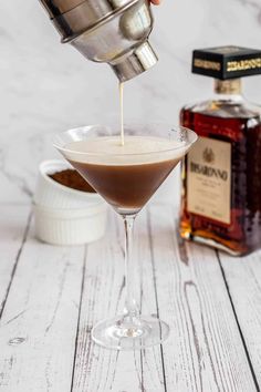 someone is pouring something into a martini glass on a white wooden table with a bottle in the background