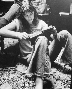 a woman sitting on the floor reading a book with two other people standing behind her