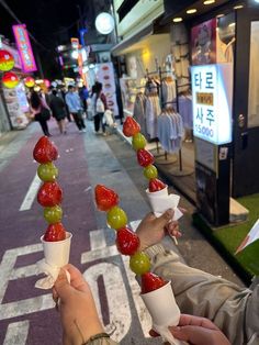 two people are holding up fruit cones on the side of the road in front of stores