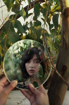 a person holding a mirror up to their face in front of a leafy tree