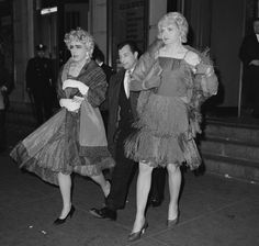 black and white photograph of two women in evening dresses walking down the street with one man