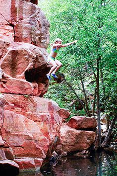 a woman climbing up the side of a red rock cliff next to a small river