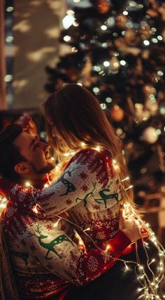 a man and woman sitting in front of a christmas tree with lights on it's chest