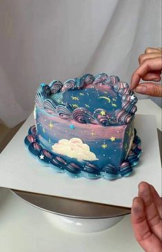 a person cutting into a blue cake with stars and clouds on it in front of a white backdrop