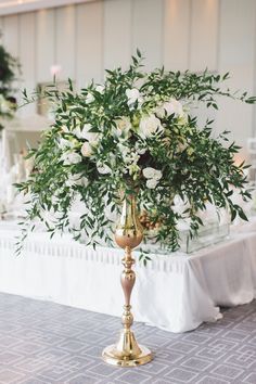 a gold vase with white flowers and greenery in it on a table at a wedding