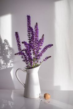 a white pitcher with purple flowers in it on a table next to a small almond