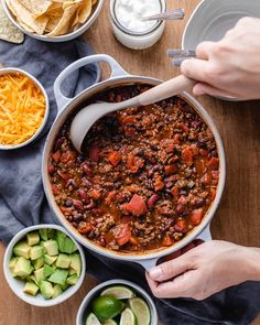 a person stirring chili into a pot with tortilla chips and avocado on the side