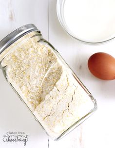 an egg and flour in a glass jar next to eggshells on a white table