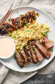 a white plate topped with meat, rice and chopsticks next to a bowl of dipping sauce