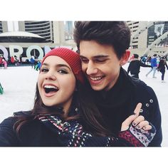 a man and woman standing next to each other in the snow with buildings behind them