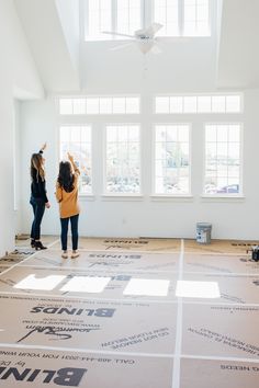 two women are standing in an empty room with windows on each side and the floor is being painted