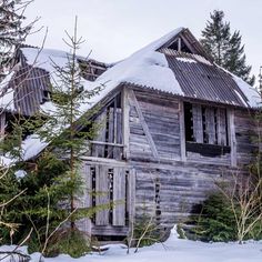 an old wooden house with snow on the roof and trees around it in front of some evergreens