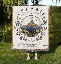 a woman standing in the grass holding a blanket with an image of mountains on it