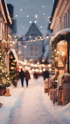 people walking down a snowy street with christmas lights on the buildings and trees in the background