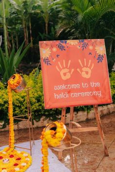 a welcome sign with flowers and decorations in the foreground is an orange banner that says welcome to the haldi ceremony