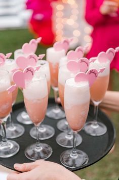 a tray filled with champagne glasses and pink hearts on top of each glass is held by someone's hand