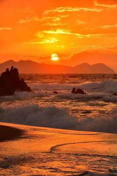 the sun is setting over the ocean with waves crashing in front of rocks and mountains