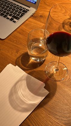 a glass of red wine next to a laptop on a wooden table with paper and pen