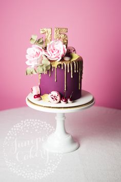 a purple cake with flowers and icing on a white pedestal against a pink background