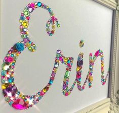 the word g is made up of colorful beads and jewels on a white background with an ornate frame