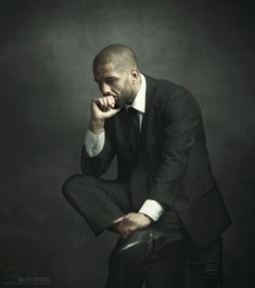 a man in a suit sitting on a stool with his hand under his chin and looking at the camera